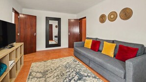 Ein Wohnzimmer mit grauem Sofa, roten und gelben Kissen, einem Flachbildfernseher, Holzregalen und einem gemusterten Teppich.