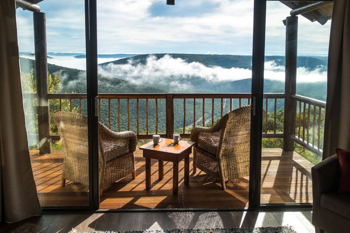 Un balcón con sillas de mimbre y una mesa, con vista a un paisaje montañoso con nubes.