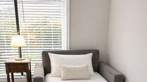 Een enkel bed met een grijs hoofdeinde en wit beddengoed in een kamer met een raam en een klein tafeltje.