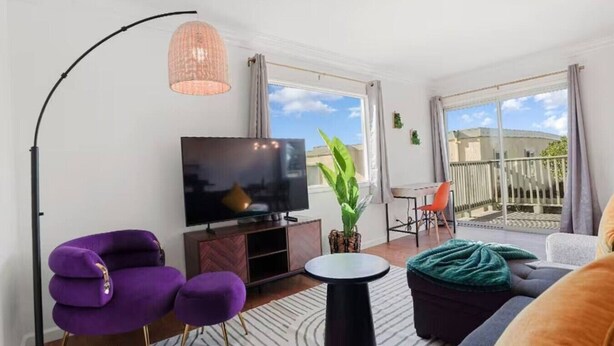 Una sala de estar moderna con un televisor de pantalla plana, muebles morados y negros, una planta grande en maceta y un balcón con vistas.