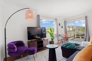 A modern living room with a flat-screen TV, purple and black furniture, a large potted plant, and a balcony with a view. - Coastal Cozy 2BR Retreat in pacifica (Pacifica)