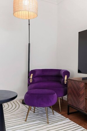 Une pièce comprenant un fauteuil violet, un pouf assorti, une table d’appoint noire, un meuble en bois et un grand téléviseur fixé au mur.