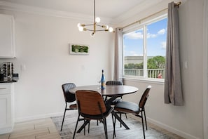 Un comedor con una mesa redonda, cuatro sillas, una araña, una ventana con cortinas y una zona de cocina con armarios y una estufa.
