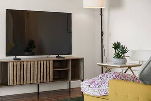 Ett modernt vardagsrum med en platt-TV på en träkonsol, en golvlampa, ett litet bord med en krukväxt och en gul soffa.