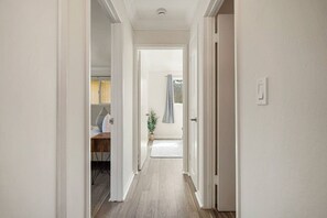 Un pasillo con piso de madera, paredes blancas y una puerta que da a una habitación con cama y ventana.