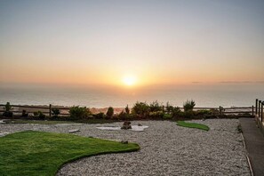 Ein Sonnenuntergangsblick von einer Terrasse mit Kiesweg, einer Rasenfläche und einem Holzzaun mit Blick auf das Meer.