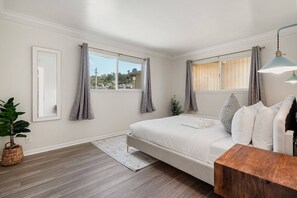 Ein Schlafzimmer mit Bett, Nachttisch, Lampe, einem Fenster mit Vorhängen, einer Pflanze und Blick ins Freie.