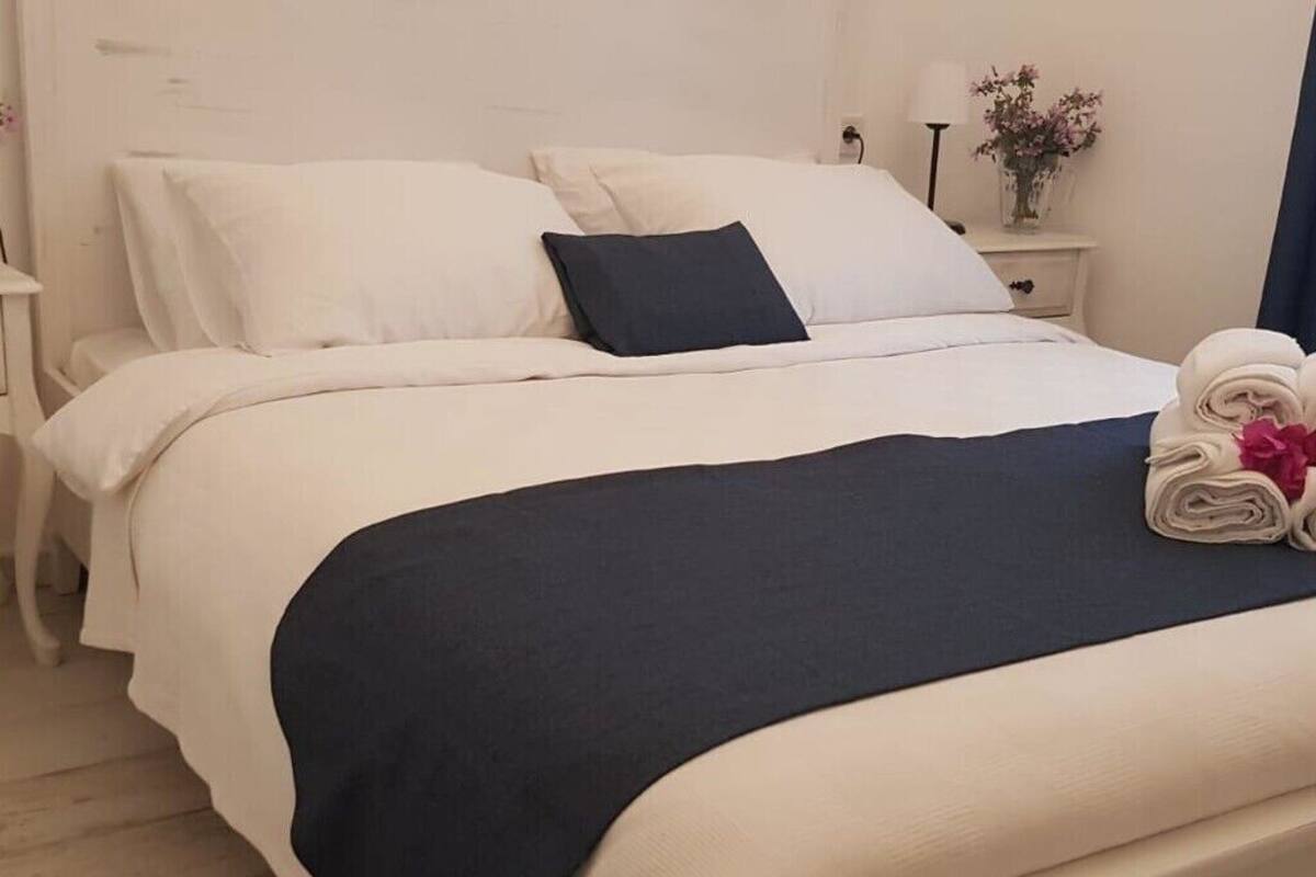 A neatly made bed with white and dark blue bedding, a nightstand with a lamp and flowers, and a rolled towel on the bed.