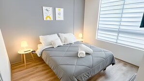 Ein ordentlich bezogenes Bett mit grauer Bettdecke, zwei Nachttischlampen und einem Fenster mit Jalousien.