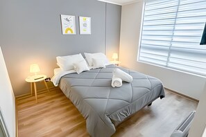 A neatly made bed with a grey duvet, two bedside lamps, and a window with blinds. - Depto acogedor cercano a metro y centro (Santiago)