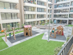 Um parquinho com balanços e estruturas de escalada, cercado por um pátio com grama sintética e um edifício com várias varandas.
