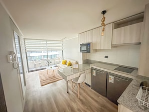 A modern kitchen with a microwave, a built-in oven, and a dishwasher. There is a dining area with a white table and chairs, and a balcony with a view. - Depto acogedor cercano a metro y centro (Santiago)
