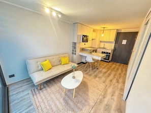 Um espaço compacto para viver, com sofá, mesa redonda pequena, cozinha compacta e porta de acesso a outro espaço.