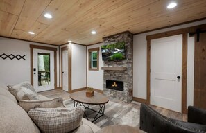 A living room with a stone fireplace, a television mounted on the wall, and a wooden ceiling. - Your Modern Creekside Retreat with Resort Perks! Modern 2bdrm + Open LoftTrundle (Ellijay)