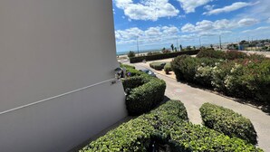 Edifici amb façana blanca, zona pavimentada amb arbustos ben podats i cel blau clar amb núvols.