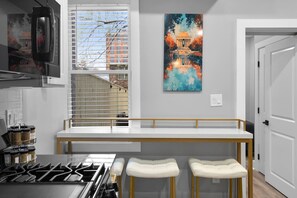 A modern kitchen with a built-in stove, a window with blinds, a white bench, and two white barstools.