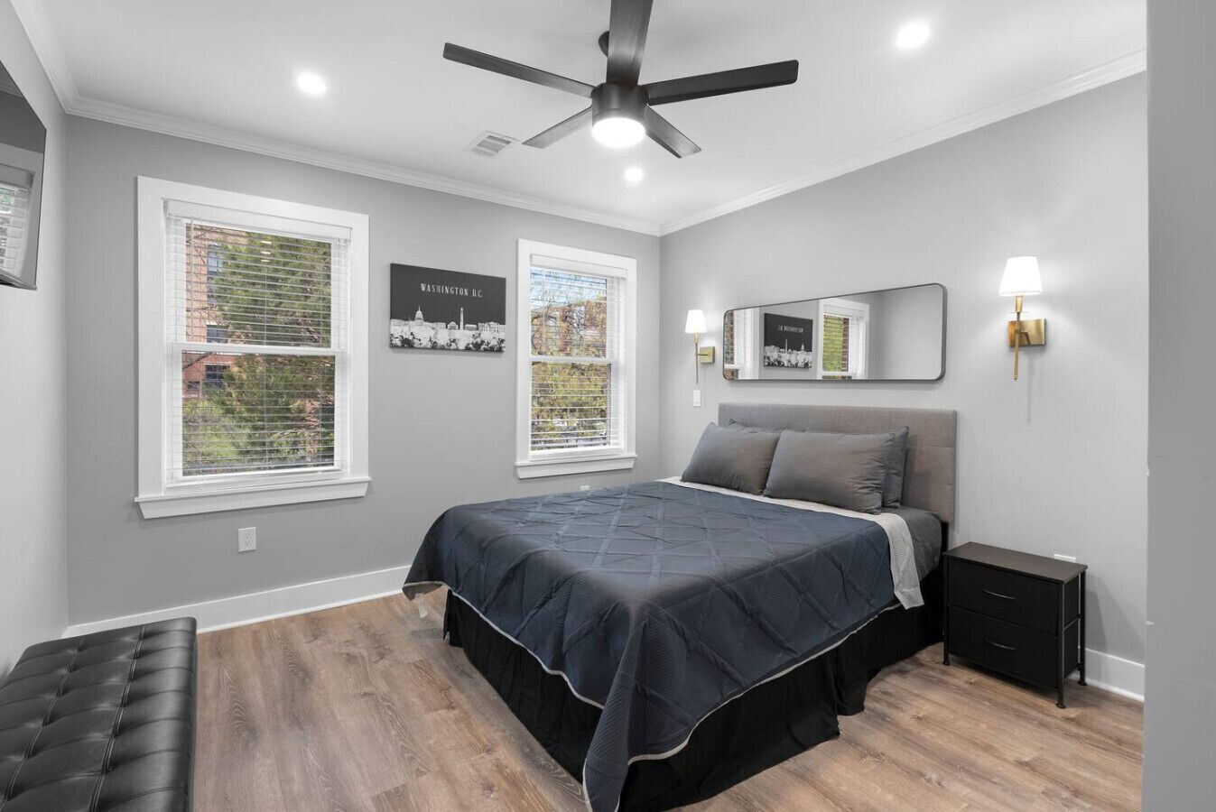 A bedroom with a bed, a nightstand, a bench, a ceiling fan, and two windows with blinds.