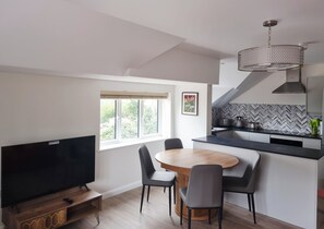 Eine moderne Küche mit Fischgrätmuster-Spritzschutz, eine Essecke mit rundem Tisch und Stühlen sowie ein Fernseher auf einem Holzgestell.