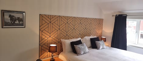 A hotel room with a geometric-patterned headboard, a bed with white bedding and patterned pillows, a bedside table with a lamp, a chair, and a framed picture on the wall.