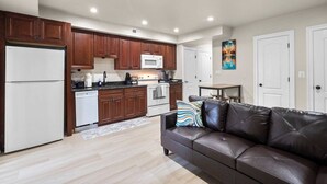 A modern kitchen with wooden cabinets, a white refrigerator, and a microwave. There is a brown leather sofa with a colorful pillow. - Modern NoMa Retreat | 2BR Near UnionMarket & Metro (Washington)