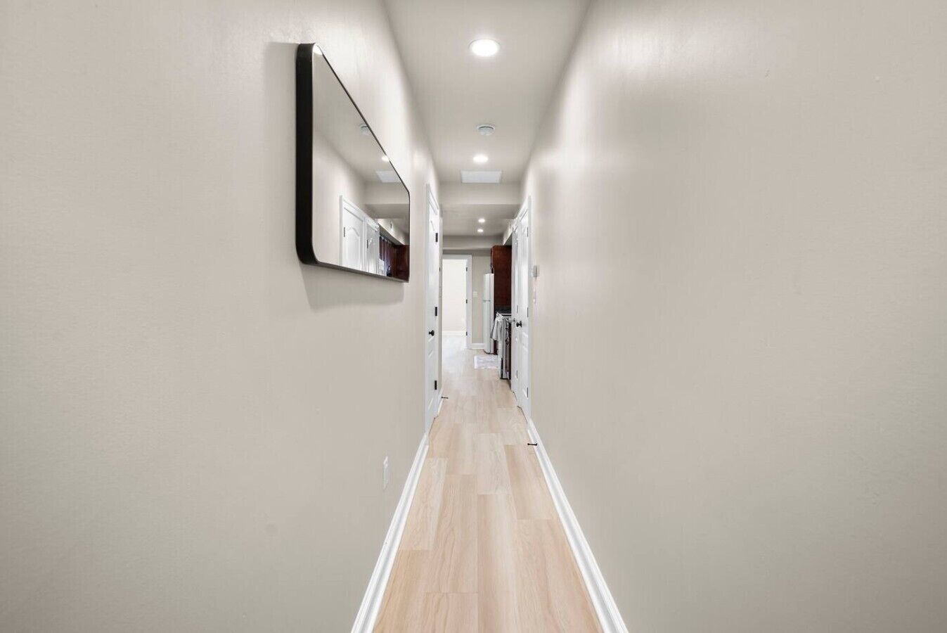 A modern hallway with a wooden floor, a large mirror on the left wall, and recessed ceiling lighting.