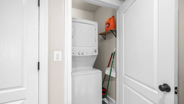 A laundry room with stacked washers and dryers, a broom, and a shelf with a detergent bottle. - Modern NoMa Retreat | 2BR Near UnionMarket & Metro (Washington)