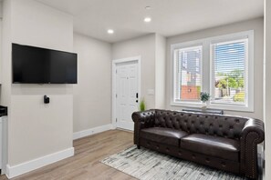 Una sala de estar con un sofá de cuero, un televisor de pantalla plana montado en la pared, una puerta y ventanas con persianas.