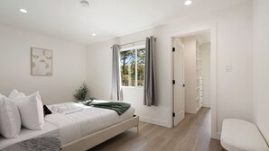 Un dormitorio con cama, mesita de noche, ventana con cortinas, una planta y un pasillo que lleva a un baño.