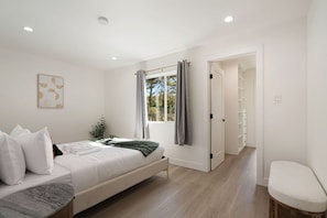 A bedroom with a bed, a bedside table, a window with curtains, a plant, and a hallway leading to a bathroom.