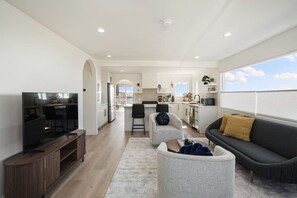 A modern living room with a TV, sofa, and coffee table, connected to a kitchen with white cabinets and a dining area.