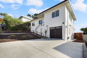 A modern two-story house with a garage, a driveway, and a landscaped area with shrubs and a small tree.
