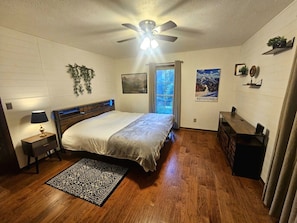 Une chambre à coucher avec un lit, un ventilateur de plafond, une table de chevet, un bureau, une fenêtre avec des rideaux et une affiche accrochée au mur.