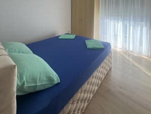 Ein Bett mit blauer Matratze, zwei Kissen, einem hölzernen Kopfteil und einem Fenster mit durchsichtigen Vorhängen.