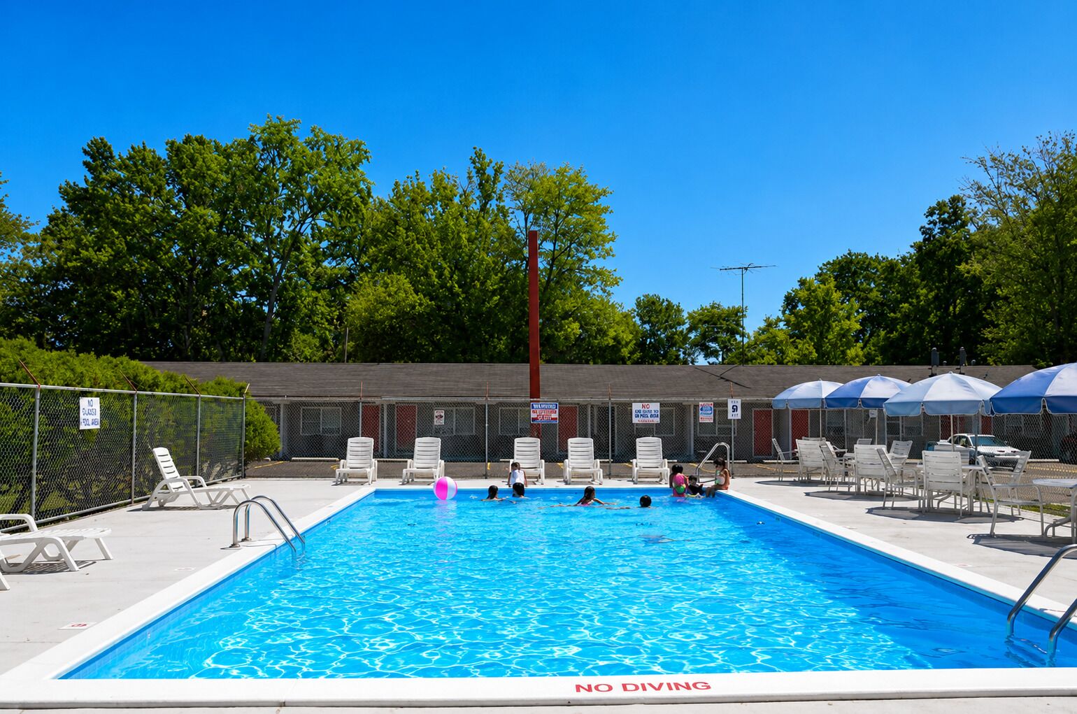 Een rechthoekig zwembad met een bordje 'NO DIVING', omringd door witte ligbedden en parasols, met een hek en bomen op de achtergrond.