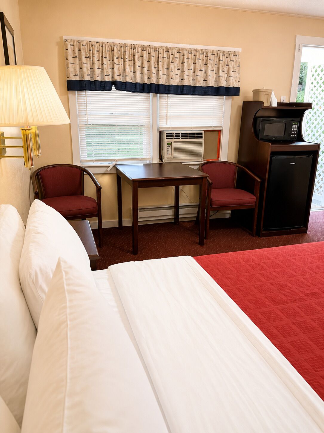 Une chambre d’hôtel comprenant un lit, une table, deux chaises, une lampe, un climatiseur, un four à micro-ondes et un réfrigérateur.