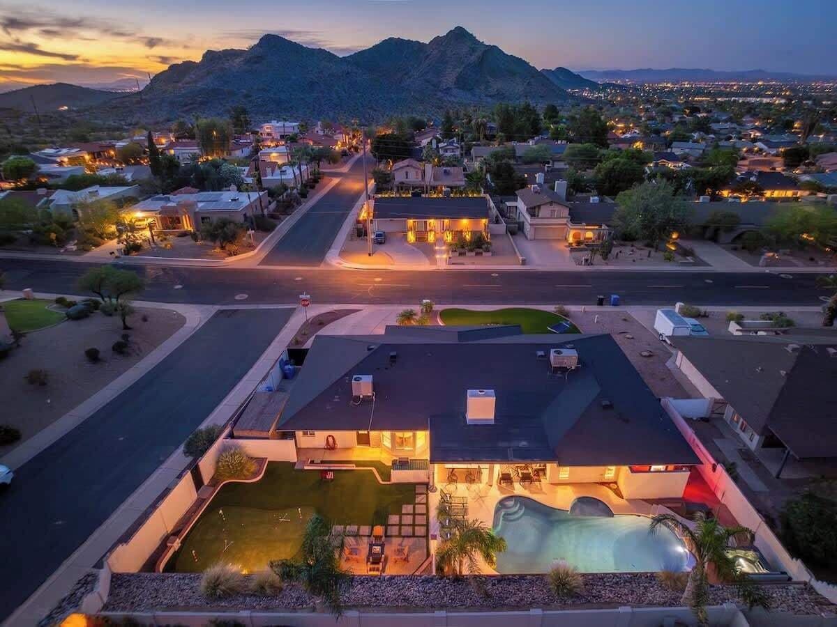 A residential area with a house featuring a swimming pool, a driveway, and a street with houses in the background.