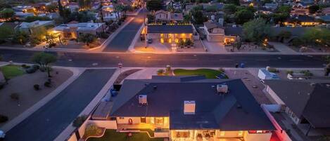 A residential area with a house featuring a swimming pool, a driveway, and a street with houses in the background.