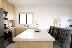 Une cuisine moderne avec des meubles en bois, un îlot central doté d’un plan de travail blanc, un four encastré et une fenêtre au-dessus de l’évier.