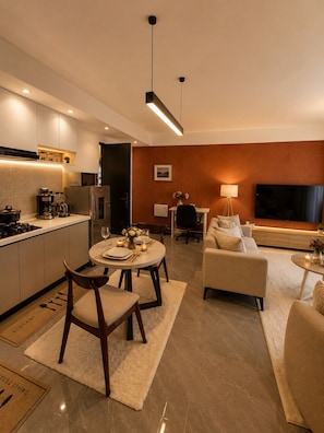 A modern living room with a sofa, a dining table with chairs, a kitchen area with a stove and refrigerator, and a TV mounted on the wall.