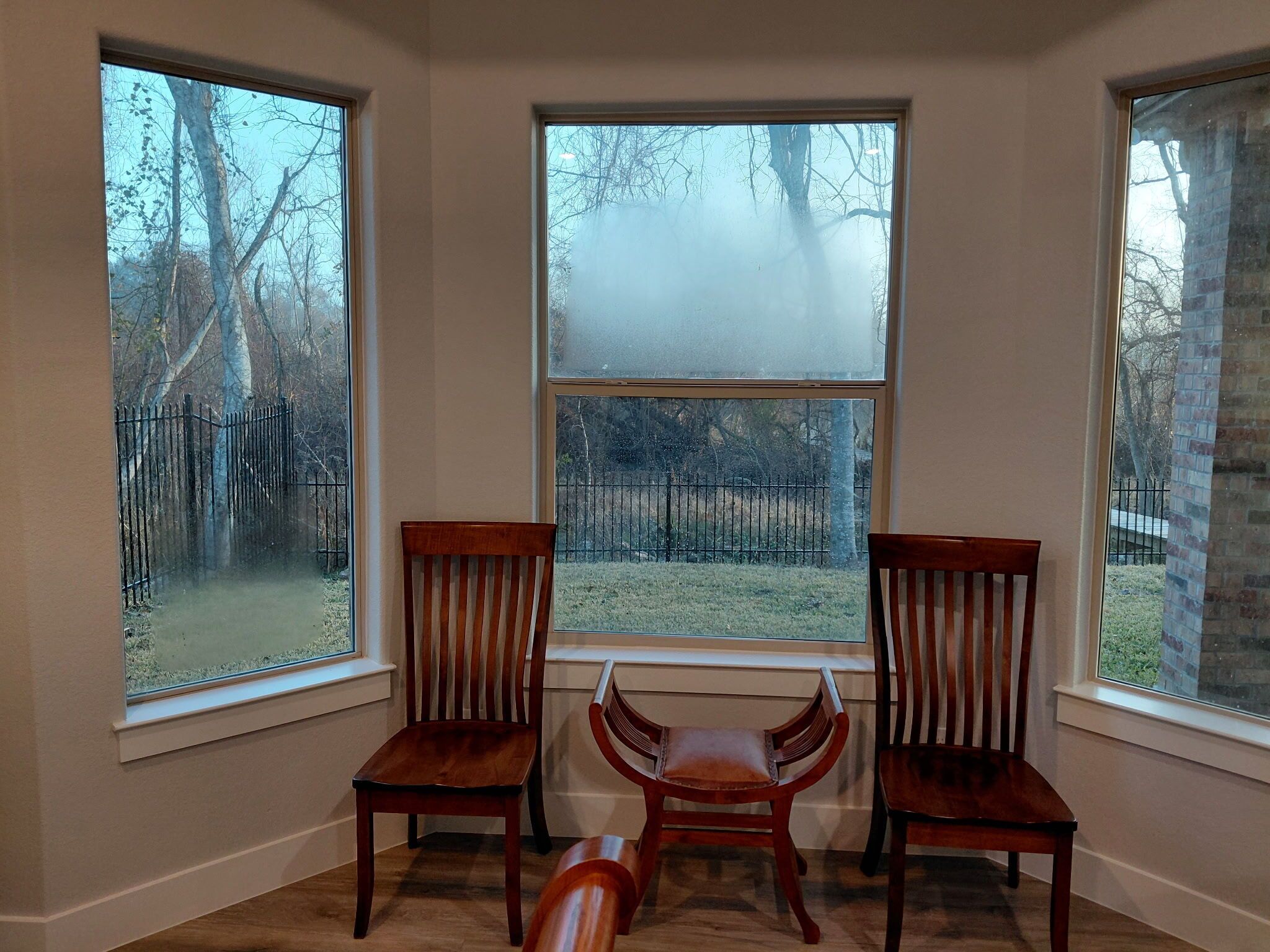 A room with three wooden chairs, a window, and a view of a fenced yard.