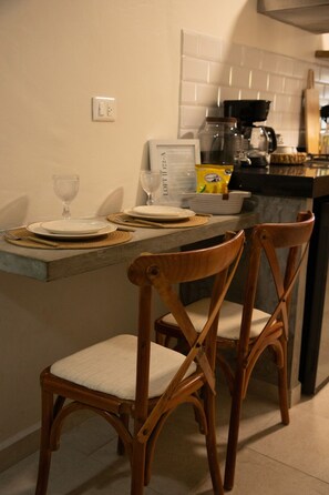 A kitchen with a dining table set for two, two wooden chairs, a coffee maker, and a framed menu on the wall.
