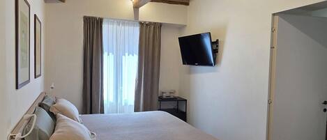 A bedroom with a bed, a flat-screen TV mounted on the wall, a window with curtains, and a door.