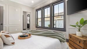 Une chambre avec une grande fenêtre, un lit recouvert d’une couverture verte, une commode en bois et une plante en pot.