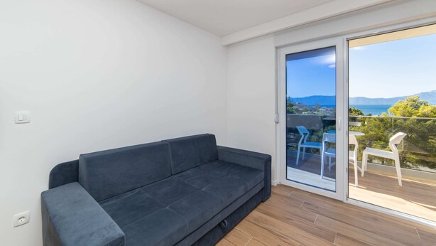 Una sala de estar moderna con un sofá gris oscuro, un ventanal corredero que da a un balcón exterior con sillas blancas.
