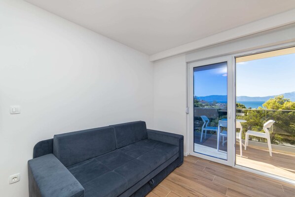 A modern living room with a dark grey sofa, a sliding glass door leading to an outdoor balcony with white chairs. - Altiora Prestige Residence 101 (Gradac)