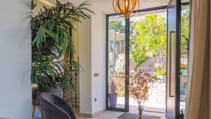 A modern room with a large potted plant, a dark armchair, a glass table with a vase, and a sliding glass door leading to an outdoor area with trees. - Villa Avril · 7 bedrooms, large pool, and unique terraces near Sitges (Olivella)