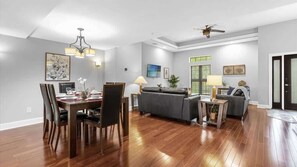 Ein geräumiges Wohnzimmer mit Holzboden, einem grauen Sofa, einem Esstisch mit Stühlen und einem Deckenventilator.