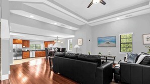 Ampla sala de estar com sofá de couro preto, piso de madeira, cozinha ao fundo e ventilador de teto.