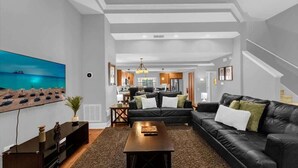 A modern living room with a black leather sofa, a coffee table, and a painting of a beach scene. - Resort 4BR Townhome | Private Pool Access & Disney Nearby (Orlando)