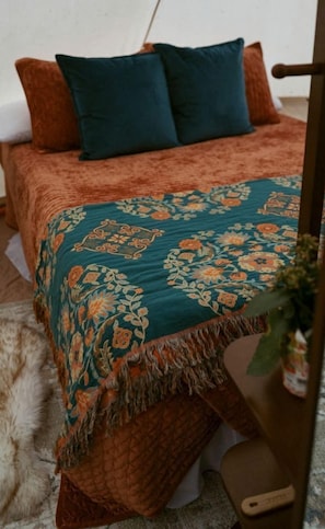 Una cama con un edredón estampado, dos almohadas y una mesita con una planta.